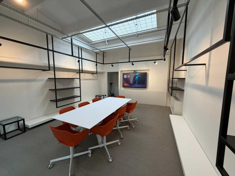 Sala riunioni dello showroom con tavolo bianco, sedie arancioni, mensole in ferro nero e lucernario